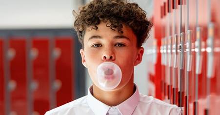 Young person blowing a bubble with chewing gum in front of school lockers.